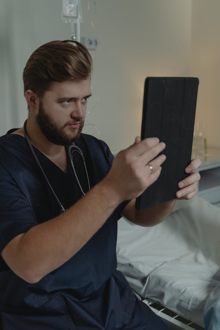 services-02 Healthcare worker with stethoscope using tablet, sitting by hospital bed, focuses on screen.